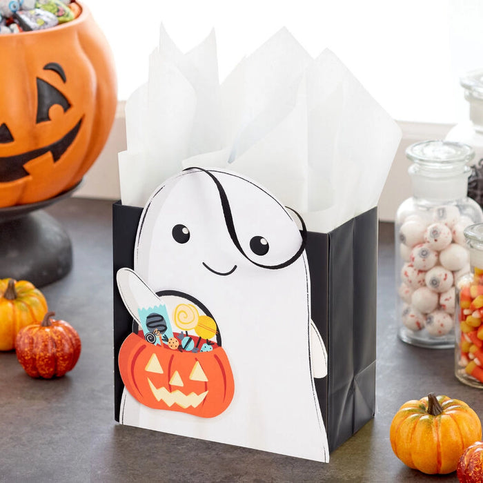 Halloween-themed gift bag with ghost design and candy on a table with pumpkins and jars.