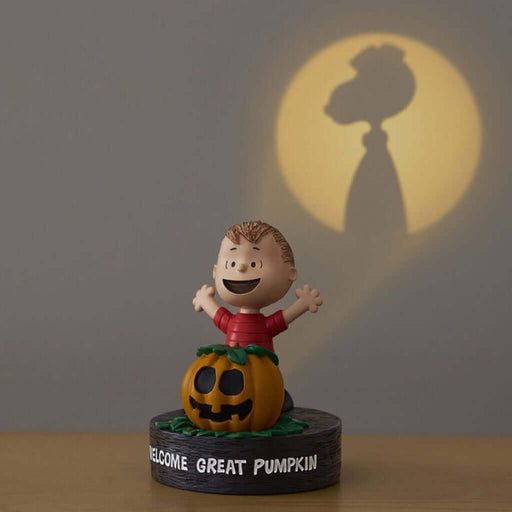Figurine of a character in a red shirt with a pumpkin, standing on a base with 'Welcome Great Pumpkin' text, against a dark background with a shadow of a rooster.
