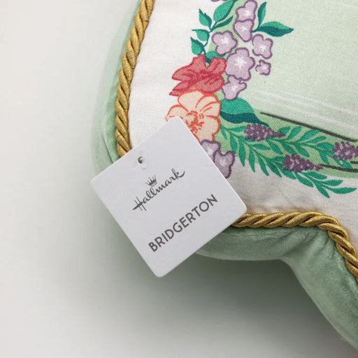 Close-up of a Bridgerton decorative pillow corner showing floral fabric, gold rope trim, and a hanging Hallmark Bridgerton product tag on a white background.