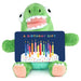 Green plush toy holding a birthday gift card with candles on a white background