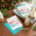 Stack of greeting cards with 'Happy Ho! Yay!' design on a wooden surface with Christmas decorations.