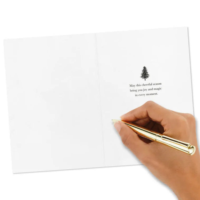 Hand holding a gold pen over a white card with a Christmas tree design and text.