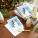 Stack of Christmas cards with a snowman design on a wooden table with decorative items.
