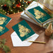Green Christmas cards with gold tree design on a wooden table with a decorated tree in the background.