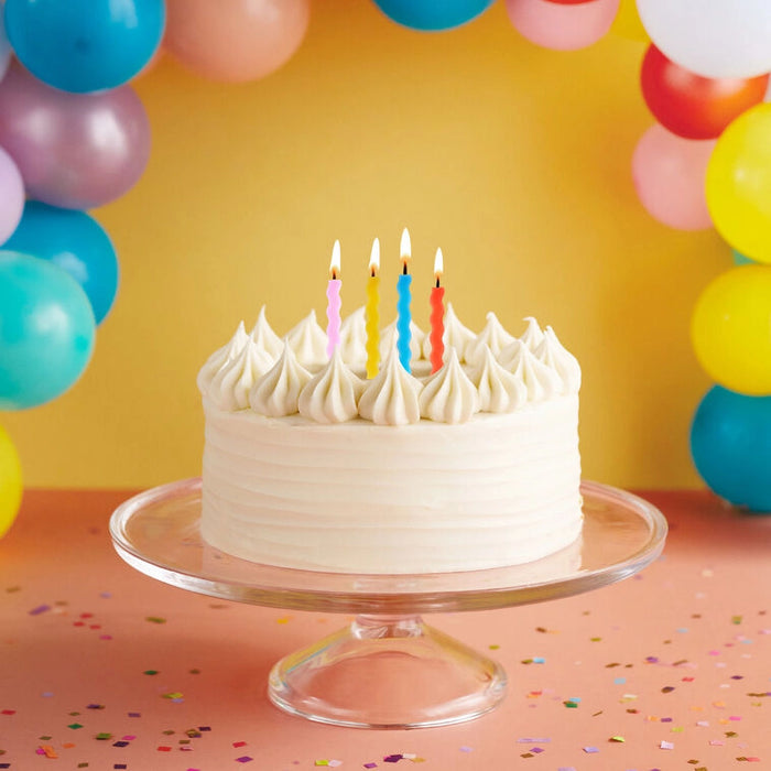 Birthday cake with lit candles on a clear stand against a colorful balloon background