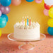 Birthday cake with lit candles on a clear stand against a colorful balloon background