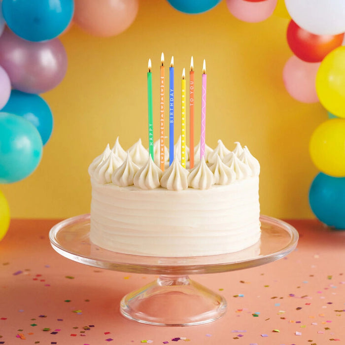 White cake with colorful candles on a clear stand against a yellow background with balloons.