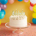 White cake with 'Celebrate' topper and colorful balloons in the background