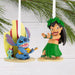 Two Disney Lilo and Stitch ornaments,  one blue character and one in a green hula outfit, hanging from a Christmas tree branch .