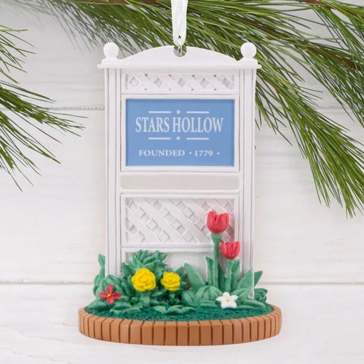 Decorative ornament with 'Stars Hollow' sign and flowers, hanging on a branch against a white background.