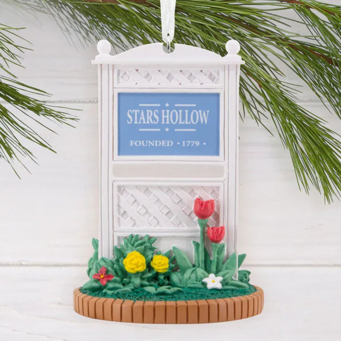Decorative ornament with 'Stars Hollow' sign and flowers, hanging on a branch against a white background.