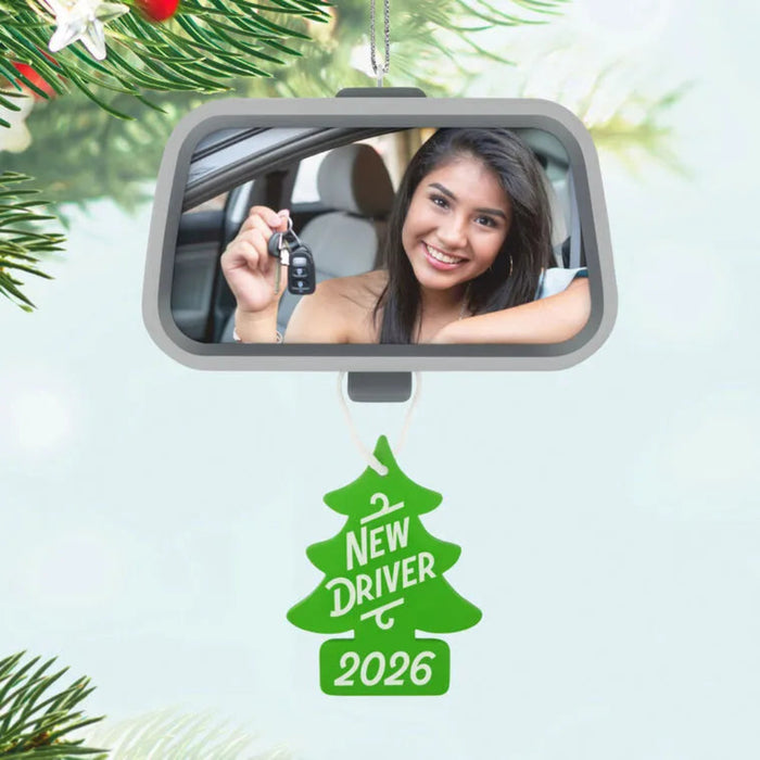 Car mirror with a reflection of a woman holding keys, Christmas tree ornament with 'New Driver 2026' text, and pine branches.