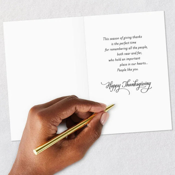 Hand holding a gold pen over a Thanksgiving card with a message on a light gray background