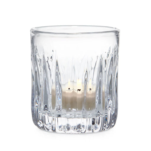 A clear glass candle holder containing two lit candles, casting a warm glow in a softly lit environment.