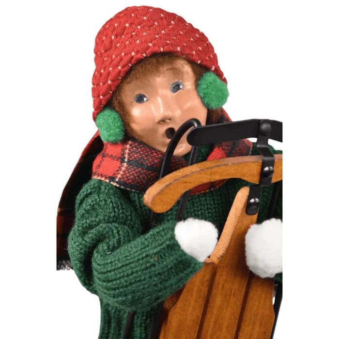 Decorative Byer's Choice  figurine of a boy in winter clothing with a red hat and green scarf, holding a  classic wooden sled.
