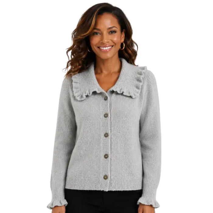 A smiling woman wearing a light gray knit cardigan with a large ruffled collar and front buttons, paired with black pants, against a plain white background.