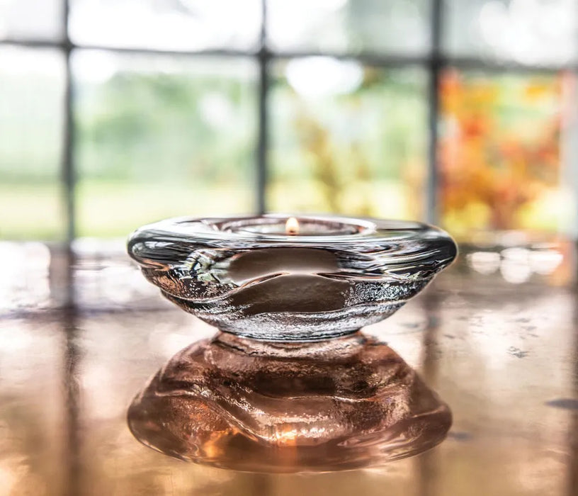 Glass  tea candle holder with a candle on a reflective surface, blurred outdoor background