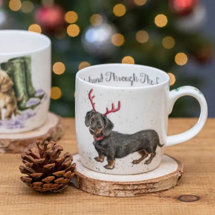 Mug with a dachshund wearing reindeer antlers on a wooden coaster with a Christmas tree in the background.