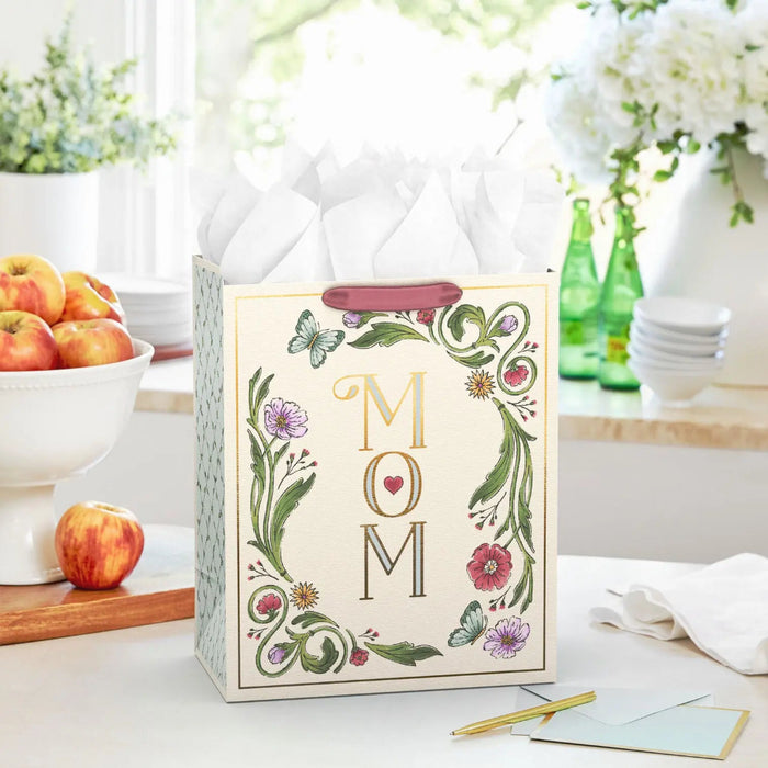 Large Mother’s Day gift bag with gold foil “Mom” lettering, floral and butterfly illustrations, and a small red heart, shown on a table with a card.