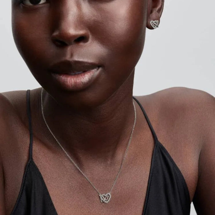 Close-up of a woman wearing a heart and butterfly shaped necklace and a earring with a neutral background