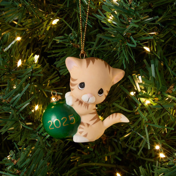 Cat-shaped ornament with a green ball labeled '2025' hanging on a Christmas tree.