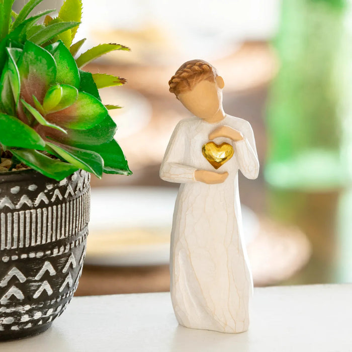 Angel figurine holding a gold heart next to a potted plant on a blurred background