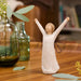 Decorative angel figurine with outstretched arms on a wooden surface with plants in the background