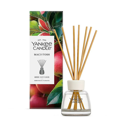 Yankee Candle Macintosh reed diffuser with a clear glass bottle, white cap, and natural bamboo reeds, shown next to its colorful apple-themed packaging box on a white background.