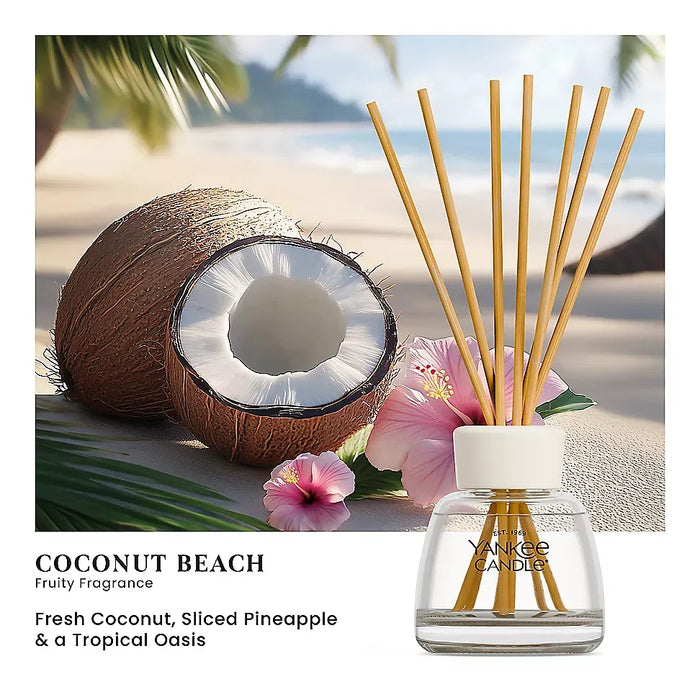 Yankee Candle Coconut Beach fruity fragrance diffuser with a glass bottle, clear liquid, white cap, and natural reed sticks, shown in front of a tropical beach scene with a coconut, pink flowers, and palm leaves. Text below reads 'COCONUT BEACH' and 'Fresh Coconut, Sliced Pineapple & a Tropical Oasis'.
