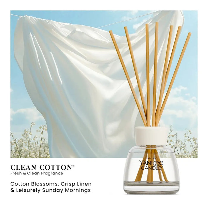 Yankee Candle Clean Cotton reed diffuser bottle with natural reed sticks, shown beside a lifestyle scene of white fabric hanging outdoors under a blue sky. Text on the image reads 'CLEAN COTTON' and 'Cotton Blossoms, Crisp Linen & Leisurely Sunday Mornings.