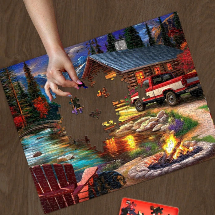 Puzzle with a scenic design of a cabin and truck, with a hand placing a piece on it.