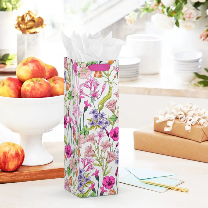 Spring floral wine gift bag with linen texture and gold‑foil tag displayed on a table beside a greeting card