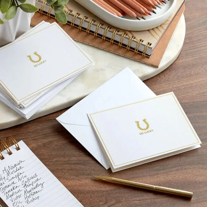 White cards with gold 'So Lucky' design on a wooden surface with stationery items.