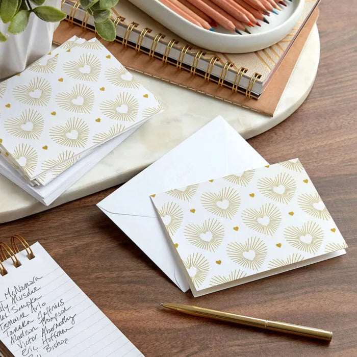 White envelopes with gold patterns on a wooden surface with a pen and notebooks.