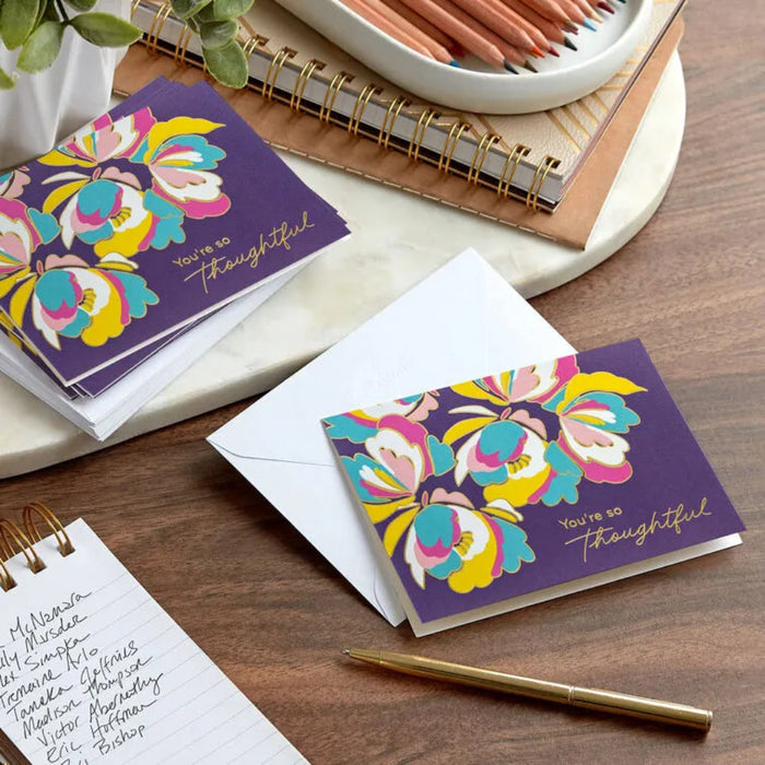 Colorful floral card with 'You're so thoughtful' message on a wooden surface with stationery items.