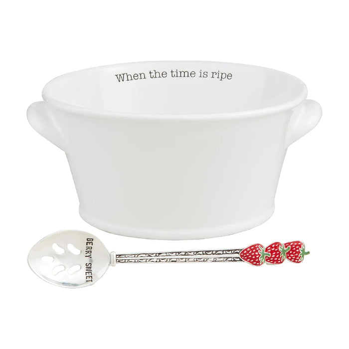 A white bowl filled with fresh strawberries beside a silver spoon on a wooden surface.