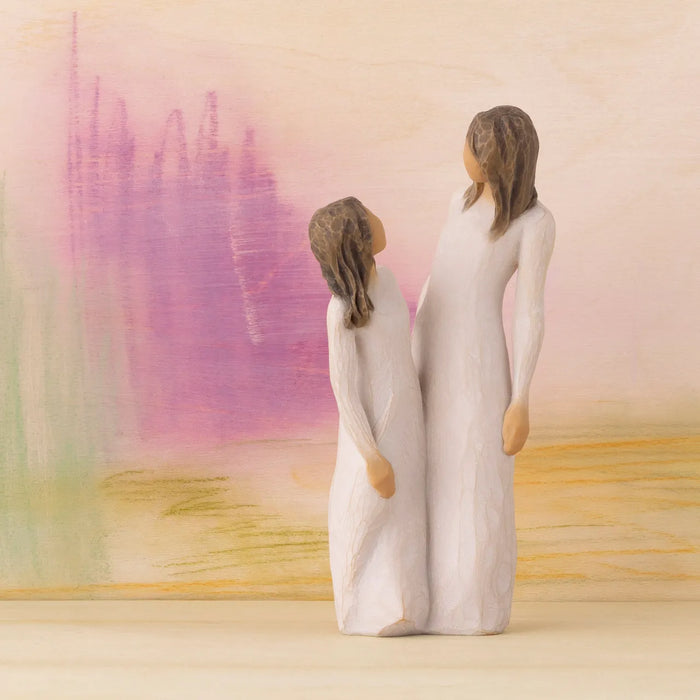 Two Willow Tree figurines of a mother and daughter in long white dresses, shown from behind against a soft pastel abstract landscape background.