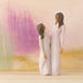 Two Willow Tree figurines of a mother and daughter in long white dresses, shown from behind against a soft pastel abstract landscape background.