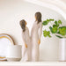 Two ceramic figures of a man and woman holding hands on a white surface with a plant in the background.