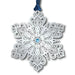 Decorative snowflake ornament with intricate patterns and a blue gem in the center, hanging from a blue ribbon on a white background.