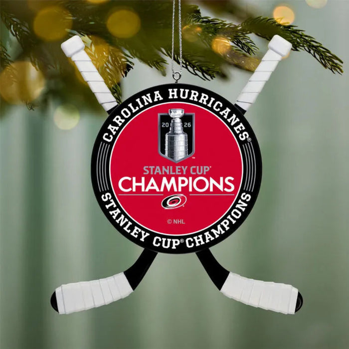 A Carolina Hurricanes Stanley Cup Champions hanging ornament shaped like crossed hockey sticks, featuring a red center with the Stanley Cup trophy graphic and white text, shown hanging from a tree branch with blurred lights in the background.