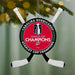 A Carolina Hurricanes Stanley Cup Champions hanging ornament shaped like crossed hockey sticks, featuring a red center with the Stanley Cup trophy graphic and white text, shown hanging from a tree branch with blurred lights in the background.