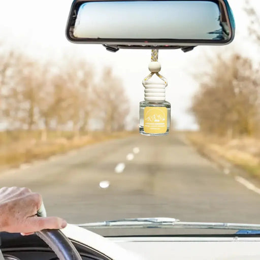A small glass car air freshener bottle with a yellow label hangs from a beaded string in front of a rearview mirror on a car dashboard. A hand is visible on the steering wheel, and the background shows a blurred road with trees.