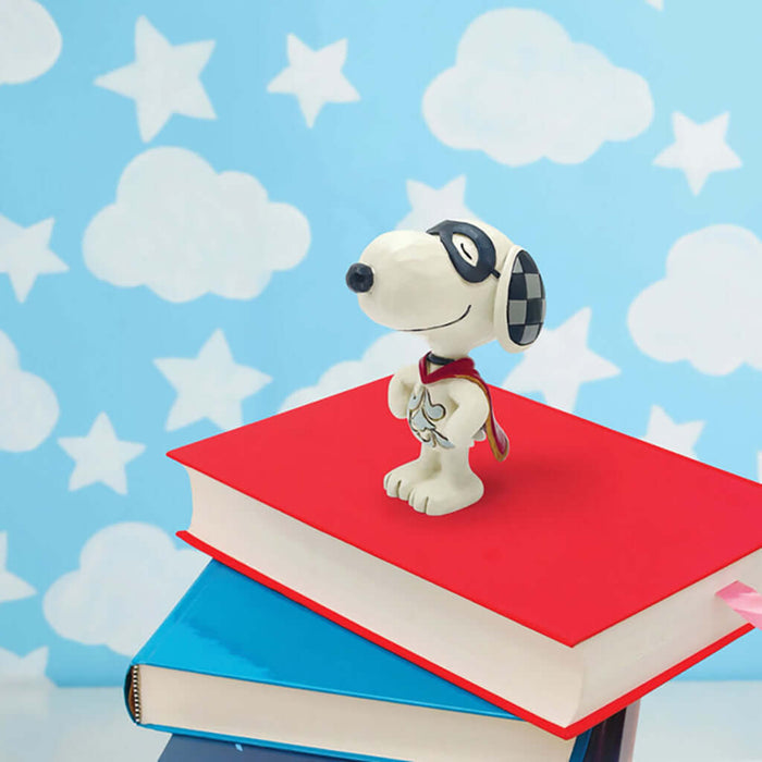 Jim Shore Peanuts mini figurine of Snoopy dressed as a superhero with a red cape and black eye mask, standing proudly on a stack of colorful books against a sky-themed background with clouds and stars