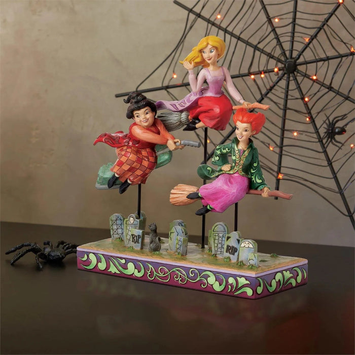 Three Sanderson Sisters  Jim Shore figurines on brooms with a graveyard background base, on a festive  Halloween background. 