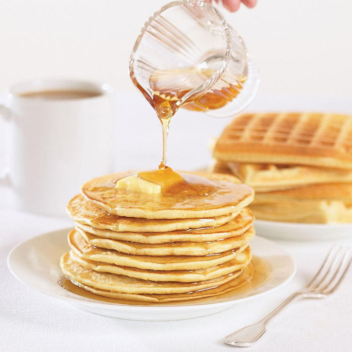 "Close-up of golden farmhouse pancakes stacked high on a white plate, topped with butter as maple syrup is poured over, with fluffy waffles and a cup of coffee in the background."