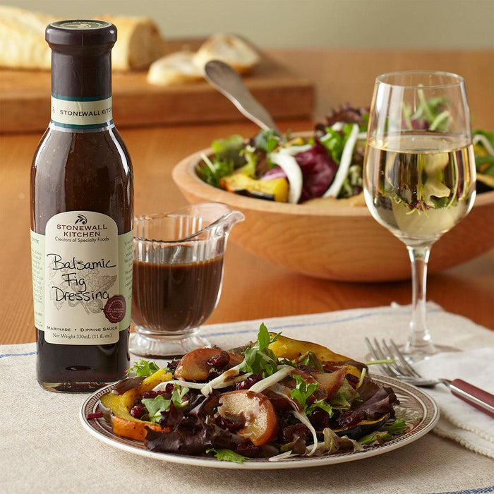 "A bottle of Stonewall Kitchen Balsamic Fig Dressing with a glass of dressed salad and a plate of food in the background."
