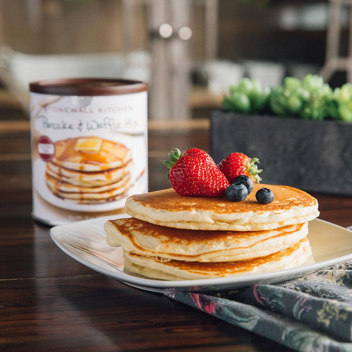 "Three golden-brown pancakes garnished with fresh strawberries and blueberries on a square plate, with a can of Stonewall Kitchen Gluten Free Pancake & Waffle Mix in the background."