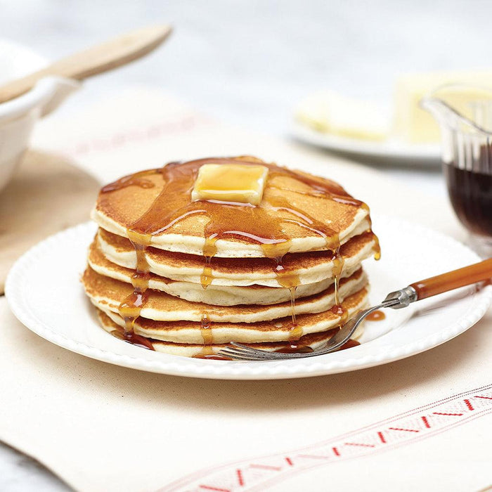 "Stack of fluffy golden pancakes topped with melting butter and drizzled with maple syrup on a white plate, made from Stonewall Kitchen Gluten Free Pancake & Waffle Mix."