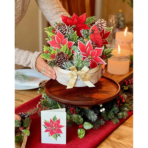 Decorative Christmas centerpiece with poinsettias and pinecones on a wooden tray, set on a table with candles and a card.